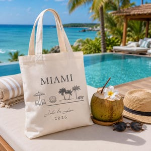 May include: A cream-colored canvas tote bag featuring the text MIAMI, John & Lisa, and 2026, along with black line art illustrations of a beach umbrella, chairs, a seashell, palm trees, and a sea turtle, printed on the front surface.