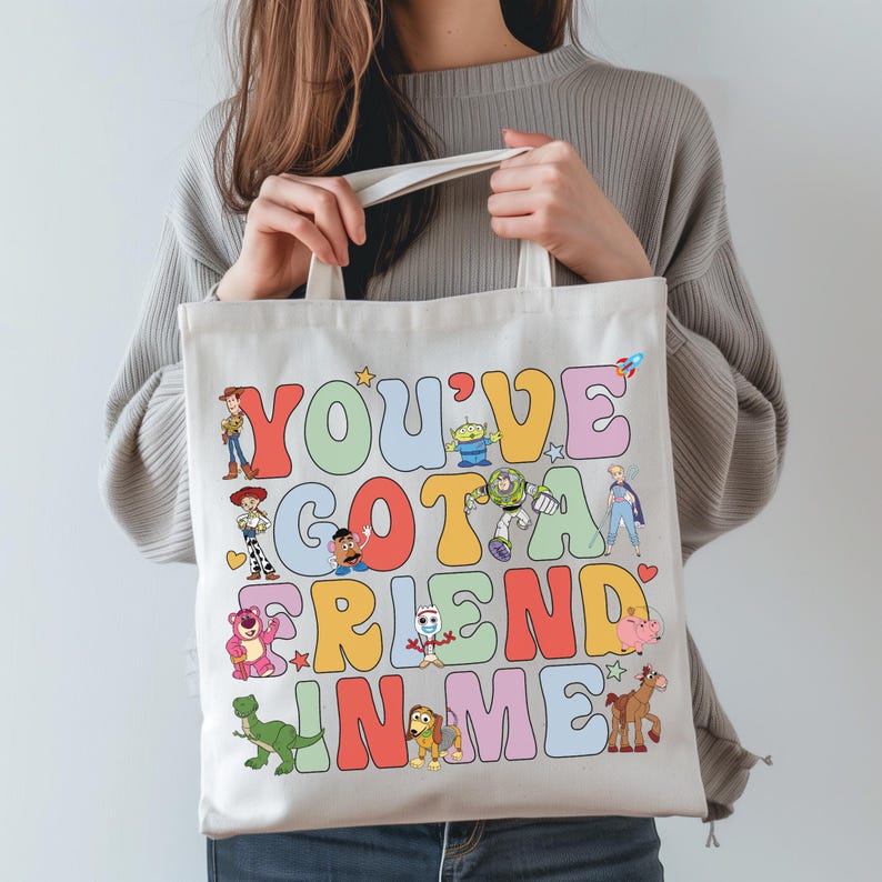 Pu&ograve; includere: Una borsa tote in tela bianca con il testo colorato "YOU'VE GOT A FRIEND IN ME" in un carattere a bolle retr&ograve;. Il design include varie illustrazioni di personaggi animati come un cowboy, un ranger spaziale, un dinosauro, un maiale e un alieno tra le lettere.