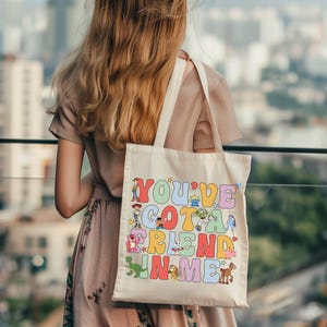 Pu&ograve; includere: Una borsa tote in tela color crema con una stampa grafica colorata che riporta il testo "YOU'VE GOT A FRIEND IN ME" in grandi lettere in stile bolla. Il design include varie illustrazioni di personaggi dei cartoni animati tra le lettere.