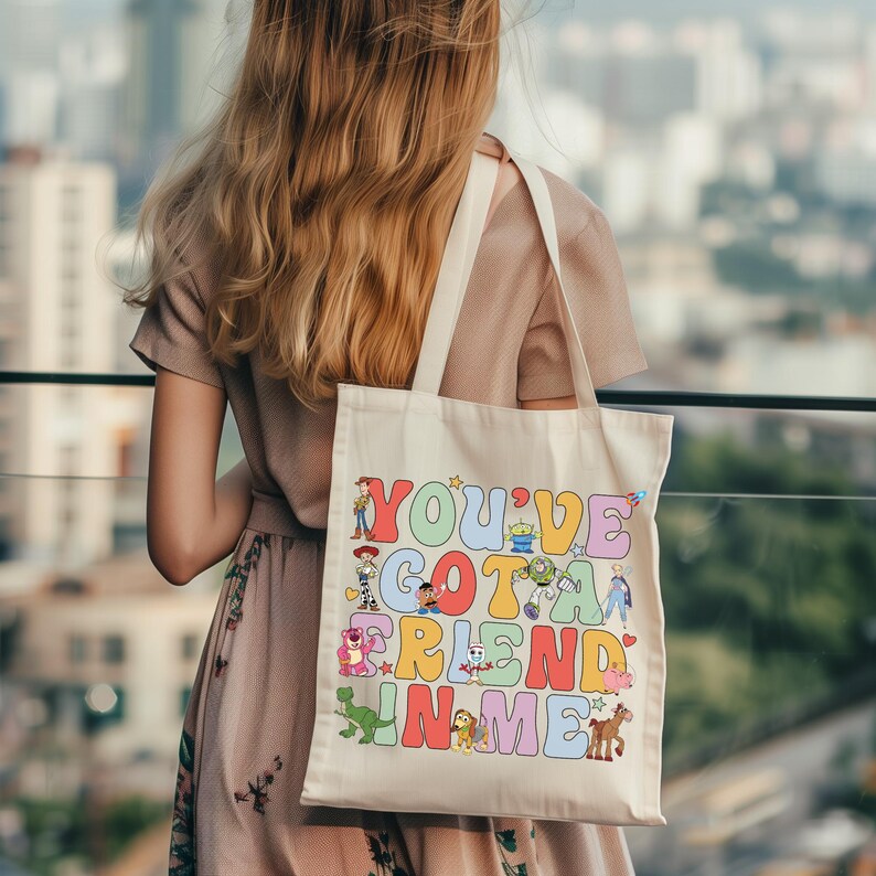Pu&ograve; includere: Una borsa tote in tela color crema con una stampa grafica colorata che riporta il testo "YOU'VE GOT A FRIEND IN ME" in grandi lettere in stile bolla. Il design include varie illustrazioni di personaggi dei cartoni animati tra le lettere.