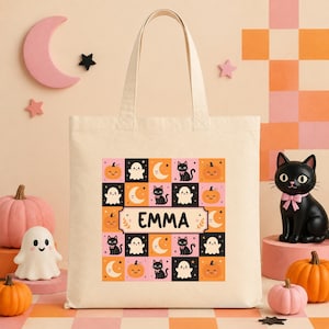 May include: A cream-colored canvas tote bag featuring a central square grid pattern of black cats, white ghosts, orange pumpkins, and yellow crescent moons. The center of the design contains a rectangular label with the name EMMA printed in black.