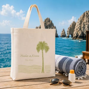 May include: A cream-colored canvas tote bag featuring a green palm tree graphic, wavy lines, and the printed text Hanhan & Kristen, Key West, and 2026. The bag has two sturdy top handles and is positioned on a wooden surface near a towel.