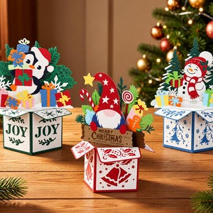 May include: Three pop-up Christmas greeting cards shaped like boxes. The left card features a penguin and gifts with the text JOY. The center card shows a gnome with a sign reading MERRY CHRISTMAS. The right card displays a snowman and gifts.