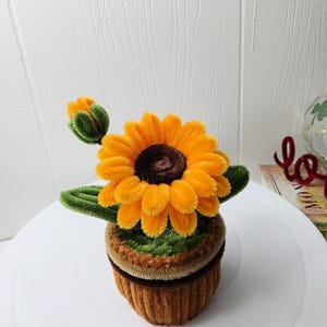 May include: A handcrafted sunflower made from fuzzy pipe cleaners, featuring bright orange petals and a dark brown center. The flower includes a small green bud and leaves, all set within a textured, brown, ribbed cylindrical pot.