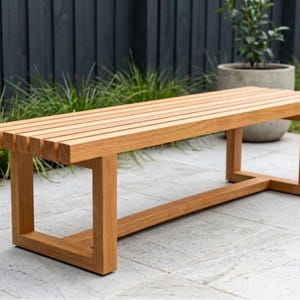 Puede incluir: Un banco rectangular de madera con un diseño de asiento de listones con tablones horizontales espaciados uniformemente. El banco está construido con cuatro patas resistentes conectadas por una barra de soporte horizontal en la base.