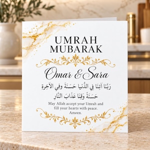 May include: A square greeting card featuring the text UMRAH MUBARAK, Omar & Sara, and Arabic calligraphy. The design includes gold marble accents and ornate gold flourishes on a white background, with the message May Allah accept your Umrah.