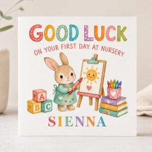 May include: A square greeting card featuring a watercolour illustration of a brown bunny in a green apron holding a red crayon. The text reads GOOD LUCK ON YOUR FIRST DAY AT NURSERY and SIENNA. Includes alphabet blocks, an easel, and books.