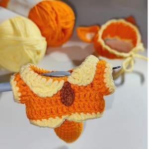 Pode incluir: Uma peça parcialmente feita de crochê nas cores laranja e amarelo com um pequeno detalhe oval marrom no peito, segurada por uma agulha de crochê prateada. Ao fundo, novelos de lã laranja e amarela e um adereço de cabeça em crochê laranja.