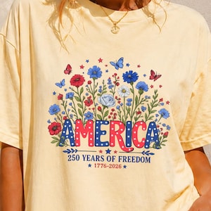 May include: A pale yellow short-sleeve t-shirt featuring a graphic of colourful wildflowers, butterflies, and the text AMERICA in red and blue with white stars. Below the graphic, the text reads 250 YEARS OF FREEDOM and 1776-2026.