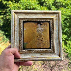 Könnte beinhalten: Ein quadratischer Kunstdruck in einem Rahmen, der ein goldfarbenes Porträt einer Person in einem gemusterten Gewand zeigt. Der Rahmen ist verziert mit einem abgenutzten Silber- und Goldfinish, einer texturierten Innenkante und einem glatten, breiten Außenrand.