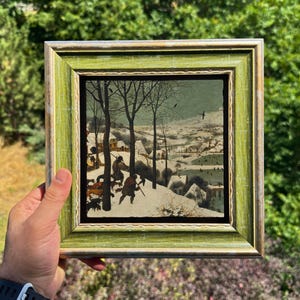Pode incluir: Uma impressão quadrada emoldurada apresentando uma pintura de paisagem de inverno com árvores nuas, caçadores e cães em primeiro plano, e uma vila coberta de neve ao longe. A obra está inserida numa moldura de madeira verde-clara com rebordo dourado.