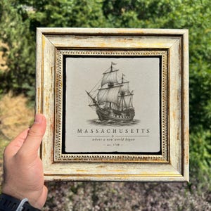 May include: A square framed wall art piece featuring a black and white illustration of a sailing ship on a light-colored tile. The text reads MASSACHUSETTS, where a new world began, est. 1788. The frame is distressed gold and off-white with ornate detailing.