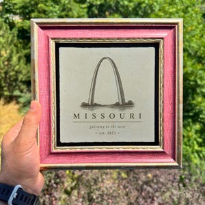 May include: A square decorative tile featuring a black line drawing of the Gateway Arch in Missouri. The text reads MISSOURI, gateway to the west, and est. 1821. The tile is encased in a textured, deep pink wooden frame with gold-toned inner trim.