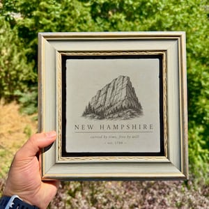 Op de afbeelding: Een vierkant ingelijst wandobject met een lichtgrijze stenen tegel voorzien van een zwarte inktillustratie van een bergtop en dennenbomen. De tekst luidt NEW HAMPSHIRE, carved by time, free by will, en est. 1788 in een schreeflettertype.