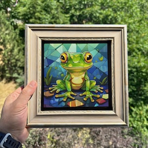 May include: A square framed wall art piece featuring a vibrant, stylized illustration of a green frog with large yellow eyes. The frog is set against a geometric, stained-glass style background of blue and green shards, encased in a light wood frame.