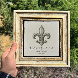 Könnte beinhalten: Ein quadratisches Wandbild in einem abgenutzten gold-weißen Rahmen mit einer zentrierten schwarzen Fleur-de-Lis-Ikone. Unter dem Symbol steht der Text LOUISIANA, soul of the bayou, und est. 1812, gedruckt auf einer hellgrauen, strukturierten Fliese.