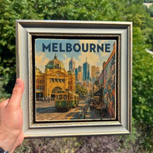 May include: A framed print featuring a cityscape of Melbourne with a yellow tram in the foreground, a historic domed building, and modern skyscrapers. The word MELBOURNE is printed in bold blue letters at the top of the artwork in a silver frame.