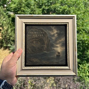 May include: A square, light-colored wooden frame containing a dark, textured print. The artwork depicts a rocky cliff face shaped like a large human eye overlooking a turbulent sea with a small sailboat at sunset. The text reads THE WEIGHT OF A NAME.