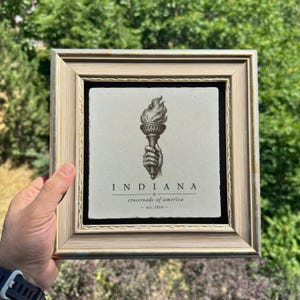 May include: A square stone tile framed in a light-colored wood frame. The tile features a black ink illustration of a hand holding a flaming torch. Below the torch, the text reads INDIANA, crossroads of america, and est. 1816.