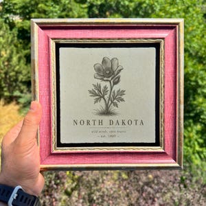 May include: A square decorative tile featuring a black line drawing of a flower centred above the text NORTH DAKOTA, wild winds, open hearts, est. 1889. The tile is encased in a textured, distressed pink wooden frame with gold-toned inner trim.