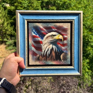 May include: A square framed artwork featuring a bald eagle with a yellow beak and white head feathers positioned in front of a waving American flag. The frame is a textured blue with gold inner trim, surrounding the central square image.