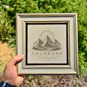 Op de afbeelding: Een vierkant ingelijst wandobject met een lichtgekleurde stenen tegel met een zwarte inktillustratie van bergtoppen en een zon. De tekst luidt COLORADO, born of stone and sky, en est. 1876. De lijst is lichtgrijs met een gouden rand.