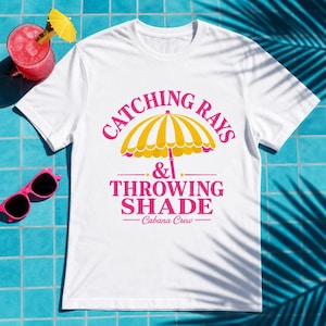 May include: A white short-sleeve t-shirt featuring a graphic of a yellow and white striped beach umbrella. The text reads CATCHING RAYS & THROWING SHADE Cabana Crew in pink lettering. A pink drink with a yellow umbrella and pink sunglasses are nearby.