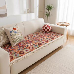 May include: A cream-colored sofa featuring a rectangular seat cover with a vibrant, multi-colored geometric pattern and cream-colored fringe trim. A square throw pillow with embroidered red and blue wildflowers rests on the sofa seat.