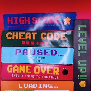 Puede incluir: Una colección de seis pegatinas rectangulares con texto y gráficos de videojuegos pixelados. Las pegatinas muestran frases como HIGH SCORE, CHEAT CODE, PAUSED, GAME OVER, LOADING y una pegatina vertical de LEVEL UP con un icono de escudo.