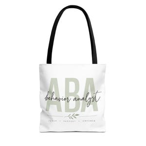 Puede incluir: Un bolso tote blanco con asas negras que presenta el texto grande en verde claro ABA, el texto en cursiva behavior analyst, un pequeño gráfico de hoja y las palabras TEACH, SUPPORT, EMPOWER impresas en negro en la parte frontal.