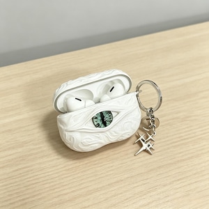 May include: A white protective case for wireless earbuds featuring a sculpted, swirling texture and a central, realistic green cat eye. The case is open, revealing white earbuds inside, and includes a silver metal keychain with star charms.