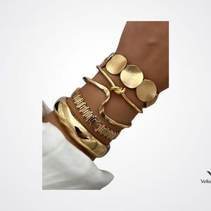 Peut inclure: Un ensemble de quatre bracelets dorés portés ensemble. La pile comprend une large manchette polie, un bracelet à maillons texturés, un jonc en fil ondulé lisse avec un nœud central et un bracelet orné de trois disques dorés plats.