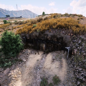 May include: A landscape view of a dirt hillside featuring a dark, recessed cave-like opening partially obscured by a green bush. The terrain is covered in dry, golden-brown grasses and rocky patches, with distant mountains under a bright, cloudy sky.