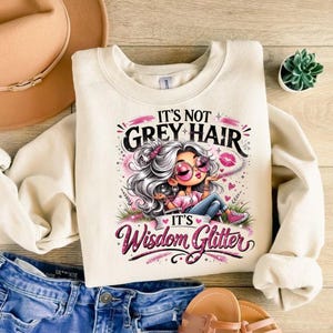 May include: A cream-colored crewneck sweatshirt featuring a graphic of a cartoon character with silver hair, pink sunglasses, and blue jeans. The text reads IT'S NOT GREY HAIR IT'S WISDOM GLITTER in black and pink script with decorative accents.