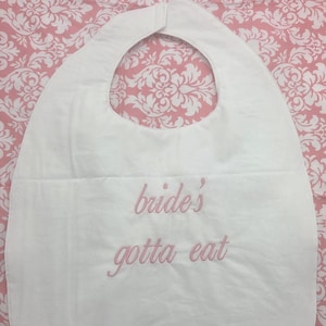 May include: White bib with a large neck opening and a pocket. The text "bride's gotta eat" is embroidered in pink thread. The bib is on a pink and white floral patterned background.
