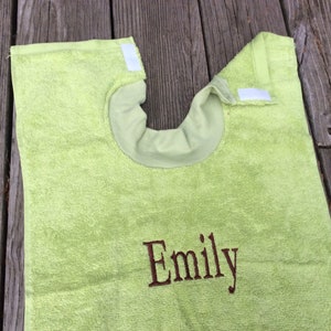 May include: A light green terry cloth bib with a neck opening and a Velcro closure. The name "Emily" is embroidered in brown thread on the front. The bib is designed to protect clothing during mealtime.