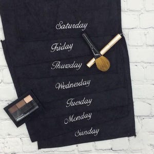 May include: A black makeup organiser with the days of the week embroidered in white. A makeup brush and eyeshadow palette are placed on the organiser, which is set against a white brick wall.