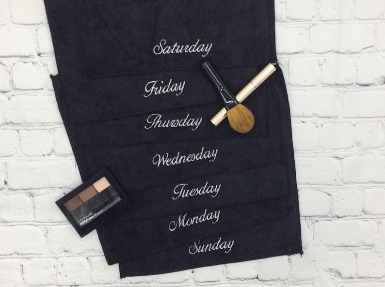 May include: A black makeup organiser with the days of the week embroidered in white. A makeup brush and eyeshadow palette are placed on the organiser, which is set against a white brick wall.