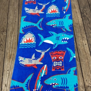 May include: A blue beach towel with a pattern of cartoon sharks in shades of blue, gray, and red. The towel also features tiki masks and surfboards. The word "Kingston" is printed on the towel.