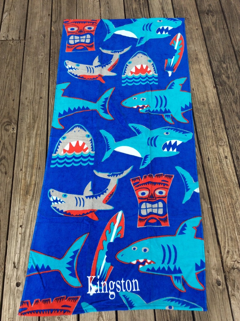 May include: A blue beach towel with a pattern of cartoon sharks in shades of blue, gray, and red. The towel also features tiki masks and surfboards. The word "Kingston" is printed on the towel.