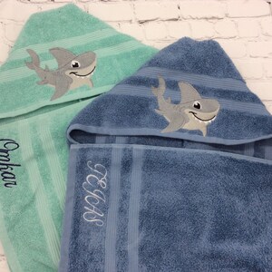 May include: Two hooded bath towels, one teal and one blue, with embroidered shark designs. The teal towel has the name "Omkar" embroidered in cursive. The blue towel has the initials "TEAS" embroidered. Both towels have striped accents.