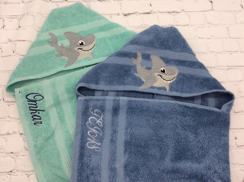 May include: Two hooded bath towels, one teal and one blue, with embroidered shark designs. The teal towel has the name "Omkar" embroidered in cursive. The blue towel has the initials "TEAS" embroidered. Both towels have striped accents.