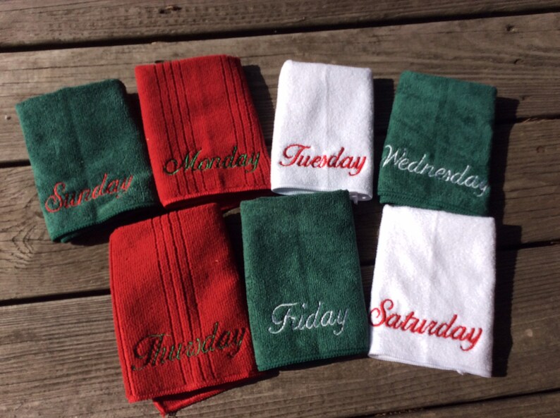 May include: A collection of seven small towels, each embroidered with a day of the week. The towels are in green, red, and white. The embroidered text is in a cursive font. The towels are arranged on a wooden surface.