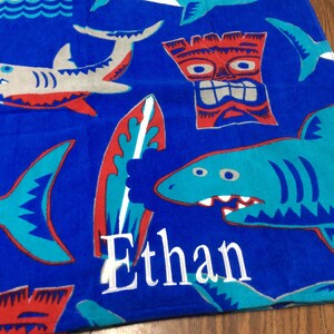 May include: Blue beach towel with shark, surfboard, and tiki designs. The towel has the name "Ethan" embroidered in white. The sharks are gray and teal with red accents. The surfboard is red and teal.