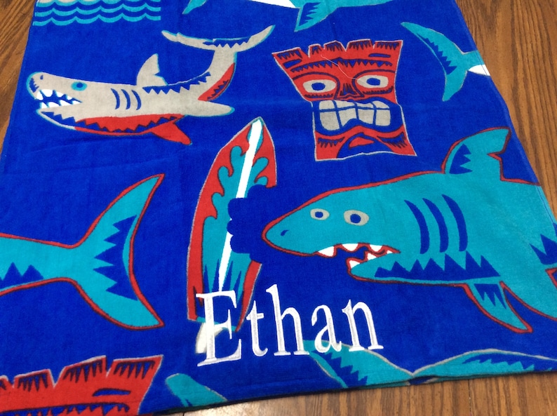 May include: Blue beach towel with shark, surfboard, and tiki designs. The towel has the name "Ethan" embroidered in white. The sharks are gray and teal with red accents. The surfboard is red and teal.