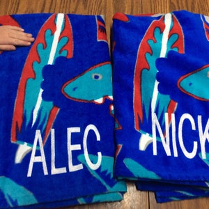 May include: Two blue beach towels with a dinosaur design in red, teal, and white. One towel is personalized with the name "ALEC" in white, and the other with "NICK".