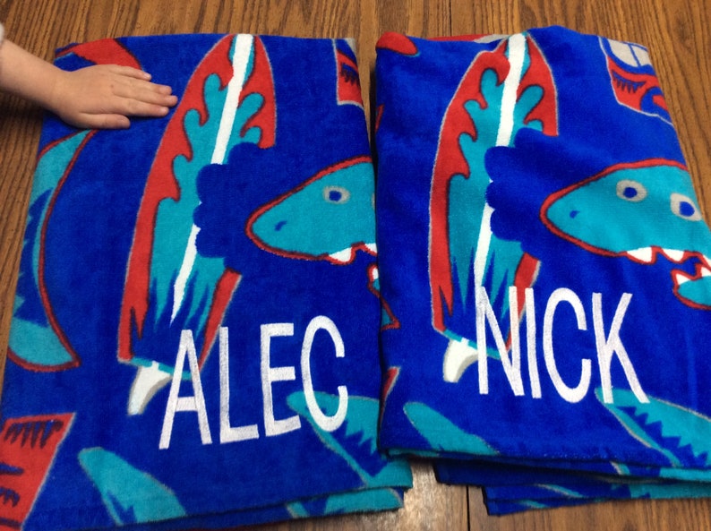 May include: Two blue beach towels with a dinosaur design in red, teal, and white. One towel is personalized with the name "ALEC" in white, and the other with "NICK".