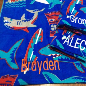 May include: Blue beach towel with shark, surfboard, and tiki designs. The towel has the names "Brayden", "Grant", and "Alec" embroidered in orange and white. The towel is folded with the names visible.