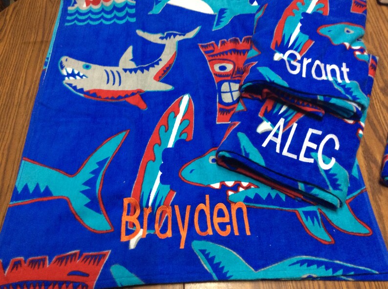 May include: Blue beach towel with shark, surfboard, and tiki designs. The towel has the names "Brayden", "Grant", and "Alec" embroidered in orange and white. The towel is folded with the names visible.