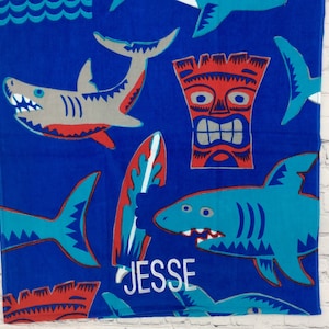 May include: Blue beach towel with shark, surfboard, and tiki designs. The towel features a shark with gray and red accents, a surfboard with red and blue accents, and a tiki with red and silver accents. The name "JESSE" is embroidered in white.